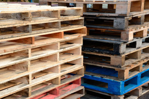 Two stacks of bare wooden pallets stacked on top of one another with one blue pallet on the bottom of the second pile